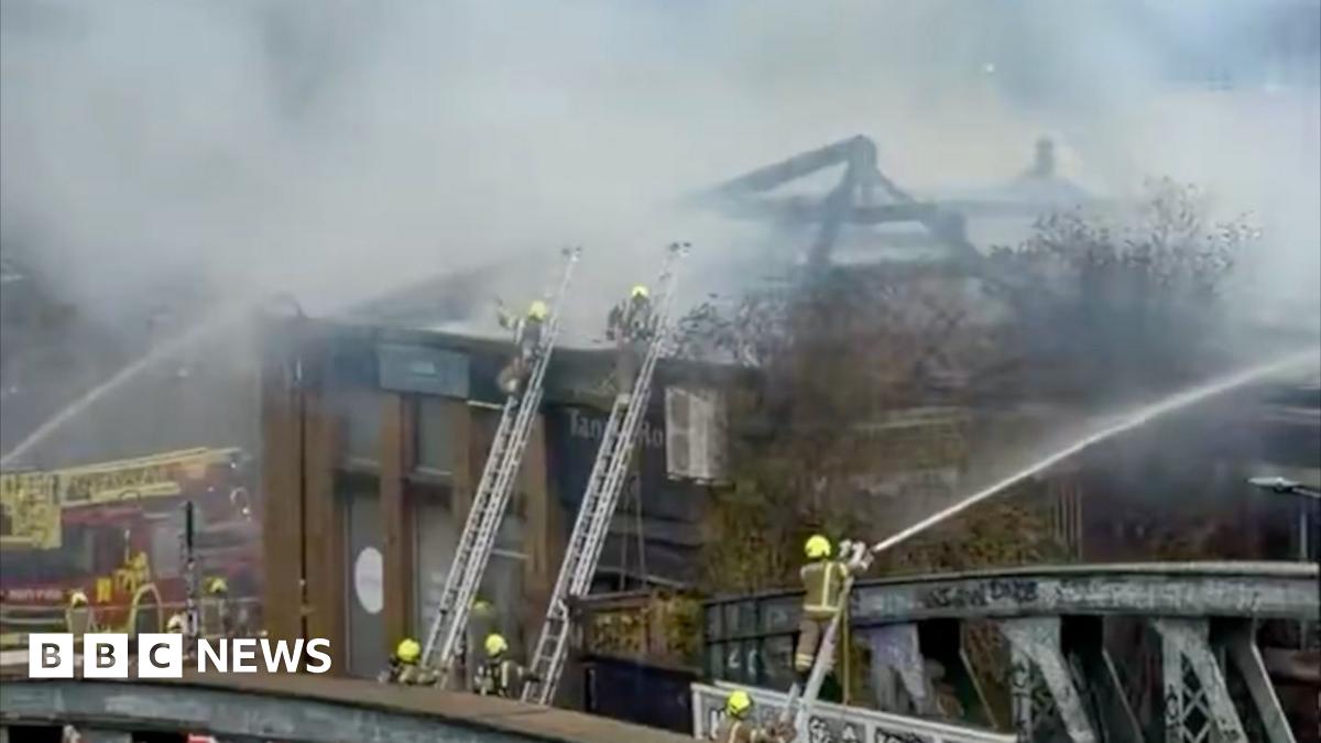 Firefighters on ladders wearing helmets spray water from hoses onto a building. A red fire engine is on the left
