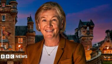 A woman wearing a brown blazer and white top. She is stood in front of a castle at sunset.