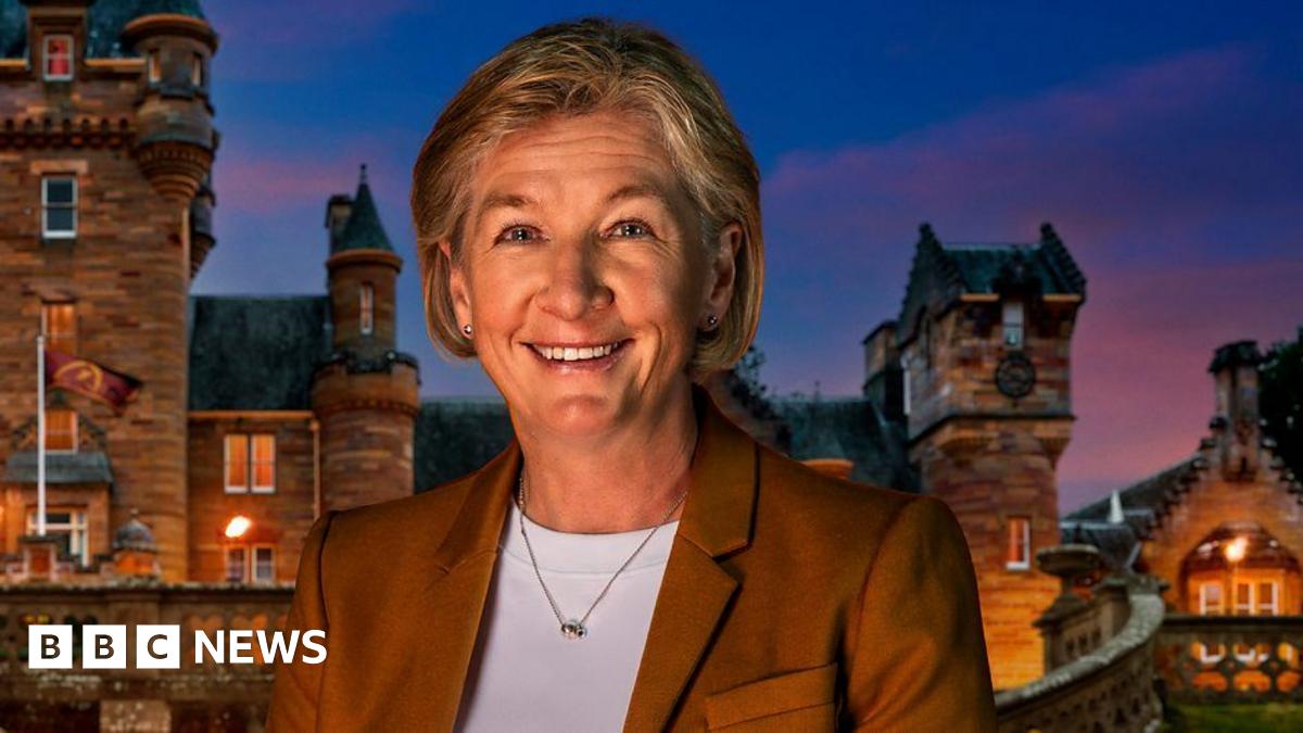 A woman wearing a brown blazer and white top. She is stood in front of a castle at sunset.