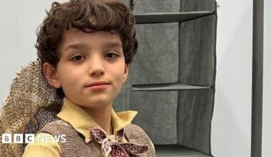 Tyne, wearing a Munchkin costume, looking at the camera. He has short dark hair, has a hat on his back, a yellow shirt, tie round his neck, and brown jacket. A rail is to his left.