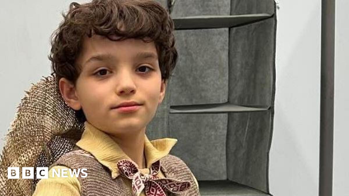 Tyne, wearing a Munchkin costume, looking at the camera. He has short dark hair, has a hat on his back, a yellow shirt, tie round his neck, and brown jacket. A rail is to his left.