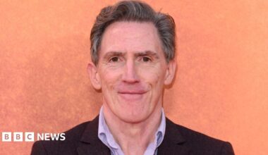 A bright orange octopus rests on a sandy beach. Its tentacles are flailing in different directions.