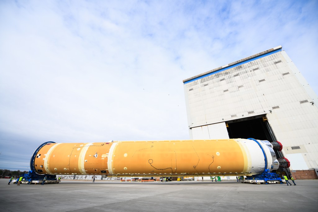 Technicians move the core stage of NASA's Space Launch System rocket--seen as a long orange cylinder.