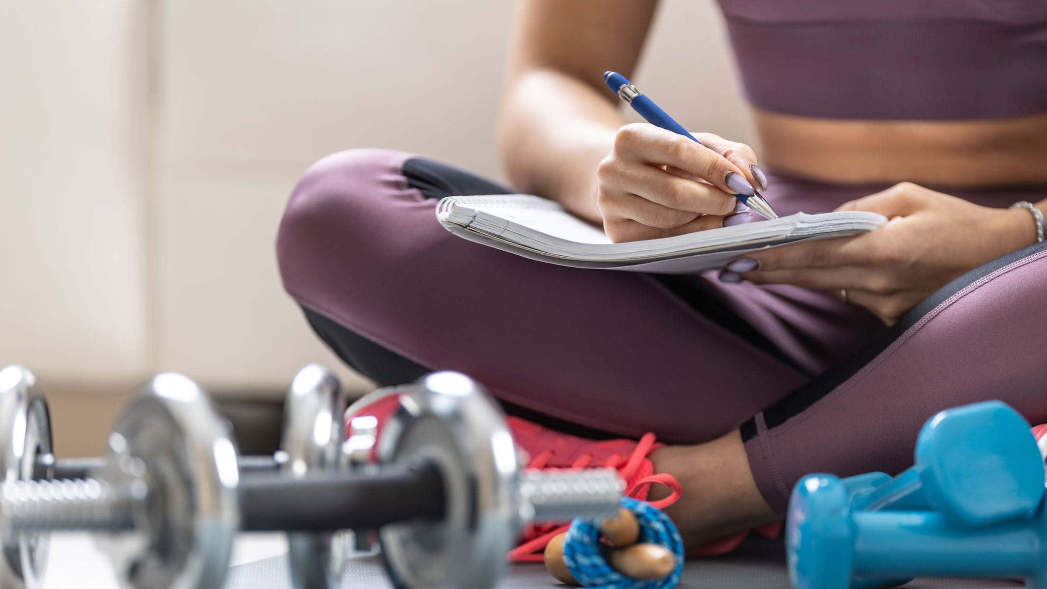 A close-up picture of a young woman writing down her fitness goals in a notebook while sitting down next to a set of dumbbells