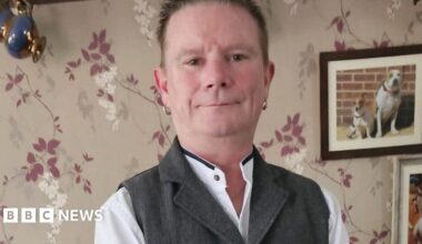 Ian Jackson-Winch. His is smiling at the camera in a living room. There is a picture of two dogs just above his left shoulder. He is smartly dressed and has an earring in each ear.