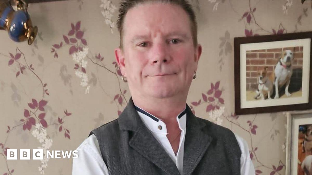 Ian Jackson-Winch. His is smiling at the camera in a living room. There is a picture of two dogs just above his left shoulder. He is smartly dressed and has an earring in each ear.