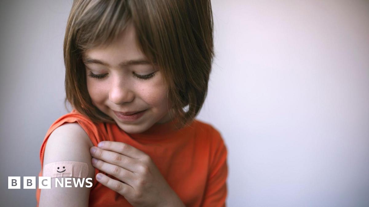 A young girl with brown, shoulder-length hair has rolled up the sleeve of her orange t-shirt to reveal a plaster with a smiley face on it on her upper arm, where an injection might go.