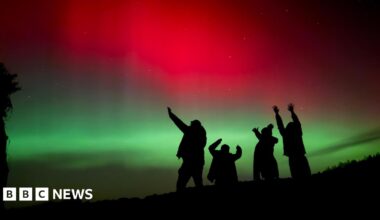 Silhouetted figures raise their arms toward the sky beneath a striking display of red and green aurora lighting the night.