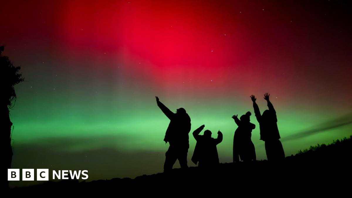 Silhouetted figures raise their arms toward the sky beneath a striking display of red and green aurora lighting the night.