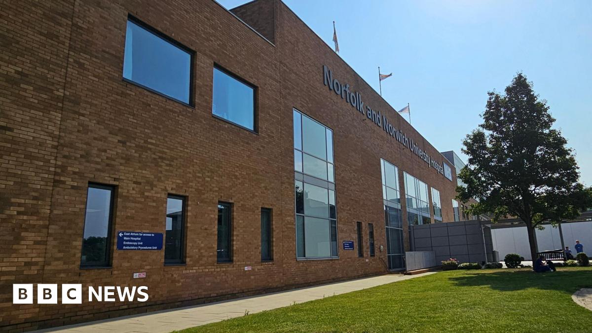 Exterior view of a hospital building. It has brown bricks and a row of tall oblong windows. In the foreground is a pavement, a tree and a grassy area. People can be seen in the distance.