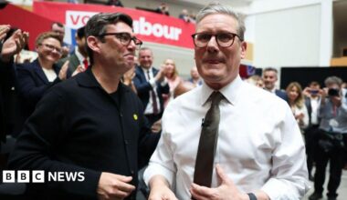Starmer and Burnham pictured together at the launch of the Labour manifesto in 2024, surrounded by an applauding crowd.