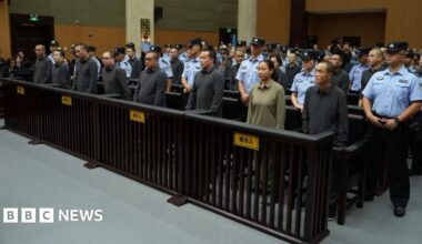 People stand in a row in a court, surrounded by security officers