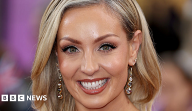 The two women stood in front of a red curtain with their arms round each other. Amy on the left is wearing all black and has shoulder-length hair with blonde highlights. Emma on the right wears a pink t-shirt with the words 'Dance to health' in white. She has tied-back blonde curly hair. They are both looking at the camera and smiling.