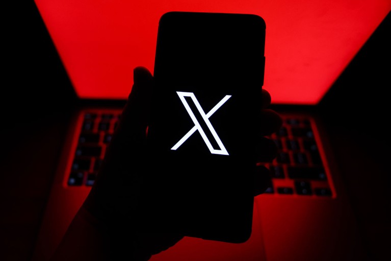 The logo for social media platform X is displayed on a mobile phone screen held up in front of an open laptop illuminated with a bright red screen.