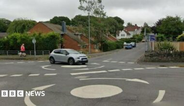 A double mini roundabout on a suburban street. Houses and parked cars can be seen. A grey car is crossing the roundabout.