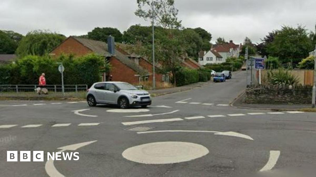 A double mini roundabout on a suburban street. Houses and parked cars can be seen. A grey car is crossing the roundabout.