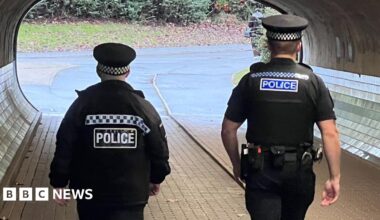 Two police officers walking through an underpass and away from the camera; they have their backs to us. Both of them are in uniform. The officer on the left is slightly shorter and is wearing a dark police officer's uniform, with a long-sleeved jacket and a peaked cap. The officer on the right is taller, is also wearing a uniform, though he is wearing a t-shirt and his bare lower arms can be seen. The pair are walking on paving stones towards the bright tunnel exit. The walls are curved, with the lower section covered with shiny tiles.