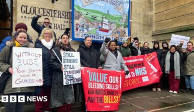 A group of striking NHS workers - perhaps around 20 people - stand in front of Gloucestershire's Shire Hall holding placards with slogans such as "Without us, your doctor would be guessing" and "Still striking, still standing, still strong".