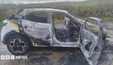 The white car completely damaged by the fire. All the windows have gone and there is debris around the vehicle.