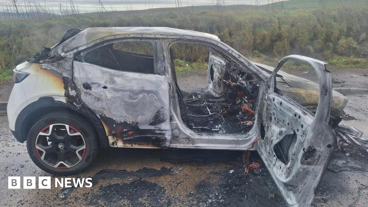 The white car completely damaged by the fire. All the windows have gone and there is debris around the vehicle.