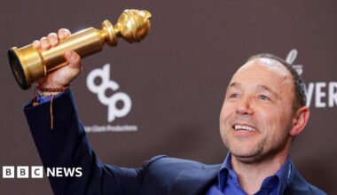 Stephen Graham has short hair and is wearing a black suit and black shirt. He is holding an Emmy award aloft with his right hand and his left hand is on his chest.