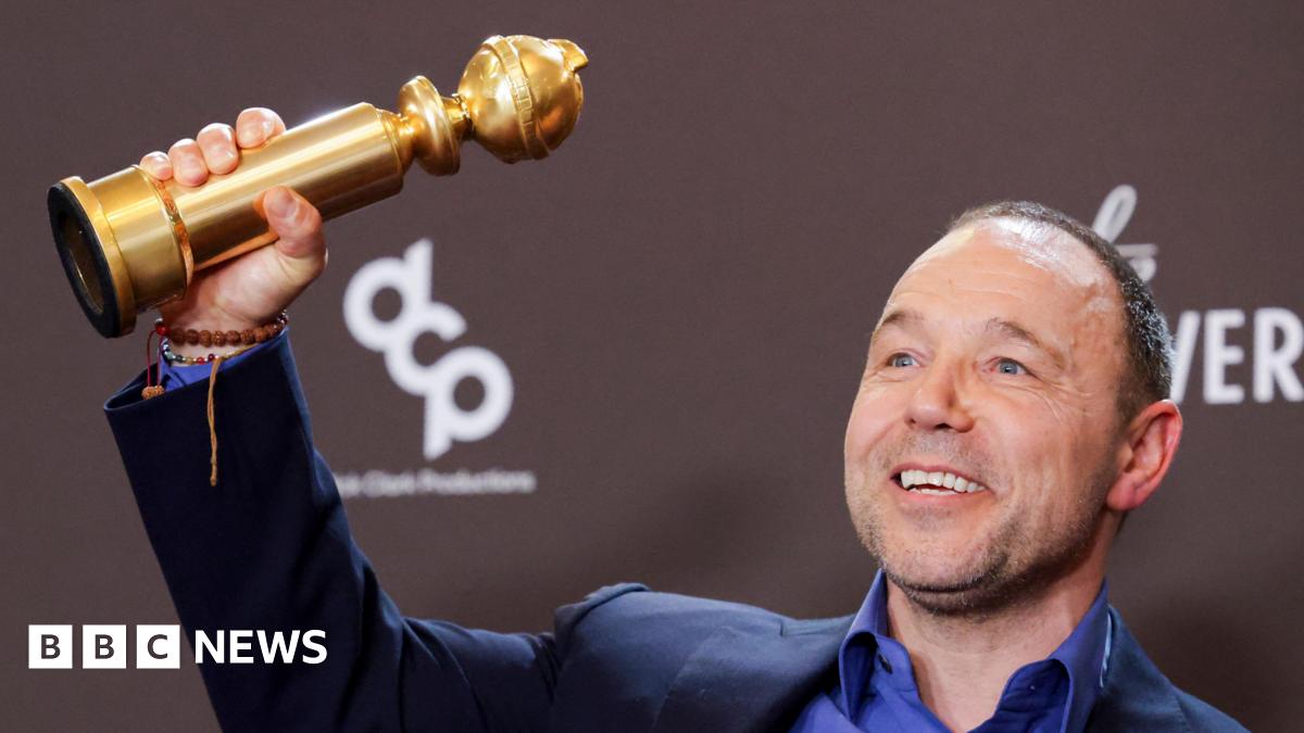 Stephen Graham has short hair and is wearing a black suit and black shirt. He is holding an Emmy award aloft with his right hand and his left hand is on his chest.