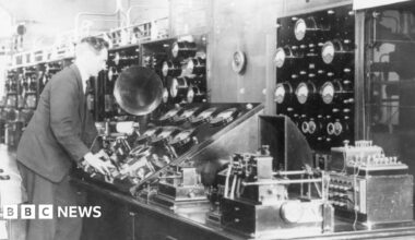A male operator stands at the main control table containing all the main control instruments including many dials and switches.  It is a black and white image and he is wearing a dark suit and tie.