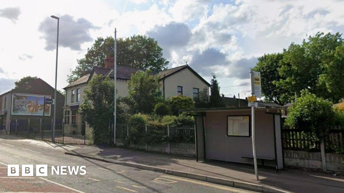 Carters Arms on Manchester Old Road Middleton is obscured by trees, a lampost and phone mast. There is a buys shelter and stop to its left.