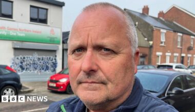 A close up image of a man with short grey hair. He is standing in front of a white building with a green banner and graffiti on the front of it. There is a row of cars parked behind him.