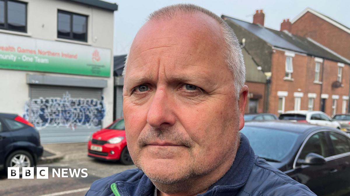 A close up image of a man with short grey hair. He is standing in front of a white building with a green banner and graffiti on the front of it. There is a row of cars parked behind him.