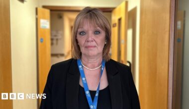 Lisa looks at the camera while standing in a corridor at the hospice. Light brown doors are on both sides of the corridor and behind her. She wears a blue lanyard with the GH white logo on it. She has a pearl necklace and a black jacket over a black top. Her hair is highlighted and shoulder length.