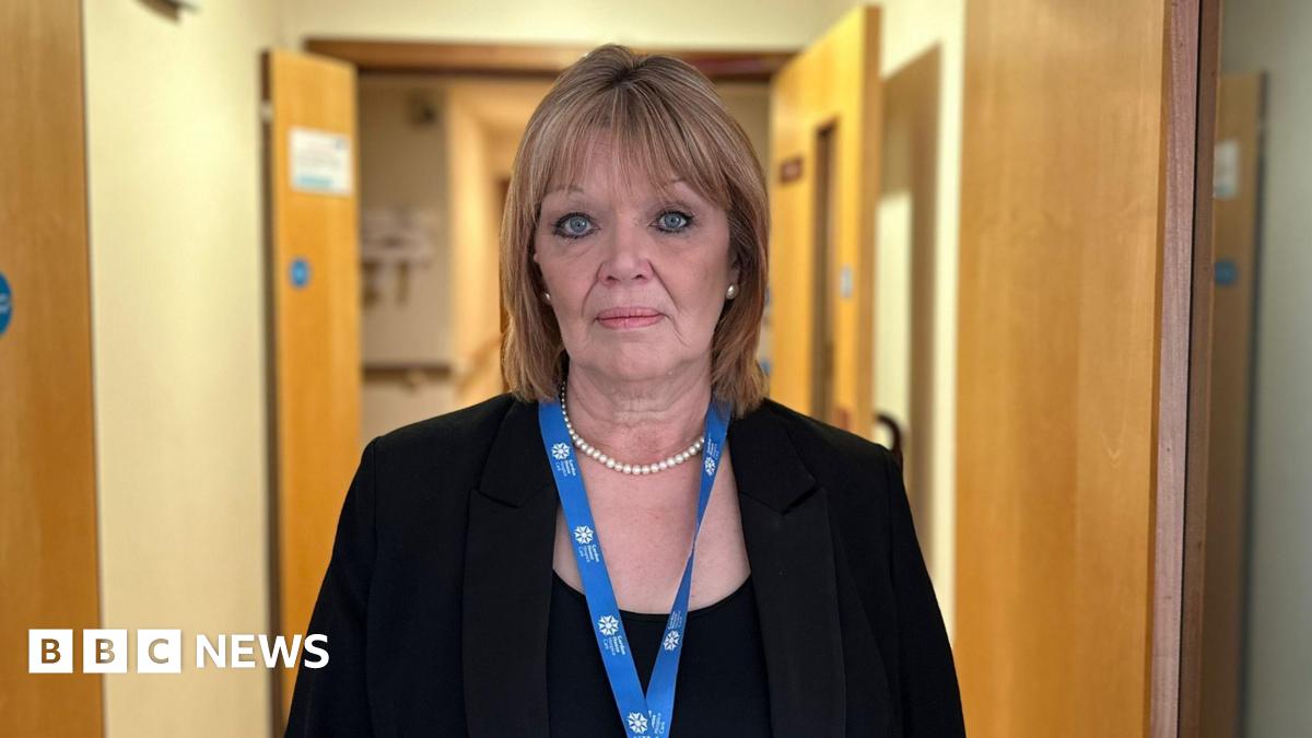 Lisa looks at the camera while standing in a corridor at the hospice. Light brown doors are on both sides of the corridor and behind her. She wears a blue lanyard with the GH white logo on it. She has a pearl necklace and a black jacket over a black top. Her hair is highlighted and shoulder length.