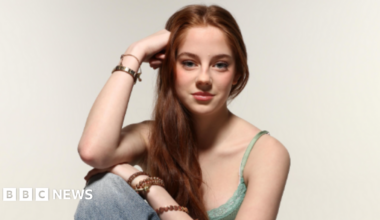 The picture shows a person seated in a relaxed pose against a plain light background. The individual has long brown hair that falls over one shoulder and is wearing a green lace-trimmed camisole top paired with light blue denim jeans. Several bracelets and bangles adorn both wrists. One arm rests on the knee while the other supports the head.