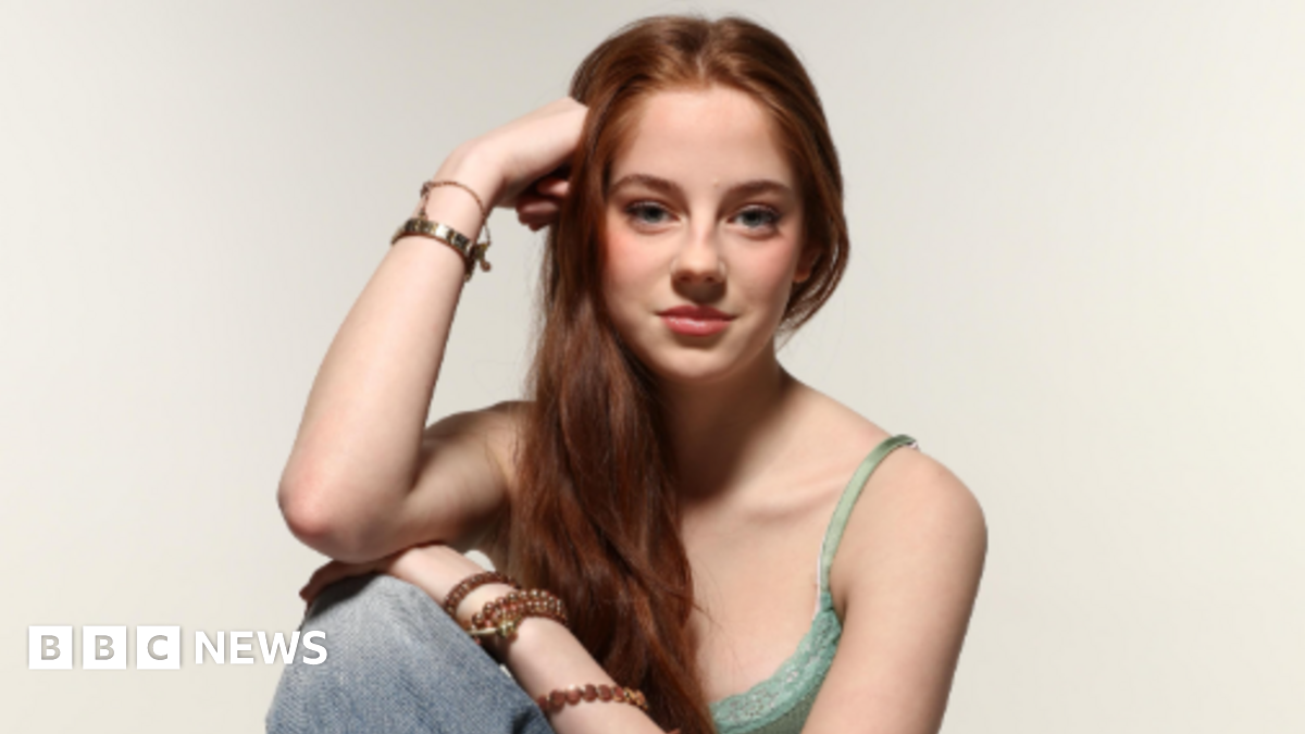 The picture shows a person seated in a relaxed pose against a plain light background. The individual has long brown hair that falls over one shoulder and is wearing a green lace-trimmed camisole top paired with light blue denim jeans. Several bracelets and bangles adorn both wrists. One arm rests on the knee while the other supports the head.