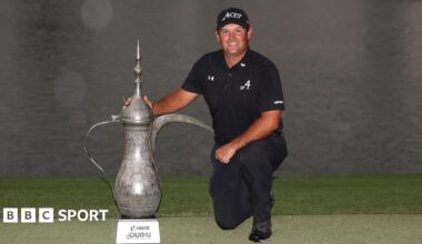 Patrick Reed, dressed in black, kneels down beside the Desert Classic trophy
