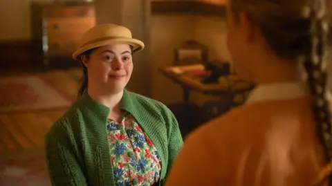A still from the TV programme, with Ellie in a hat, floral dress and green cardigan. She is facing another person, seen from behind, who is out of focus and in the foreground of the shot