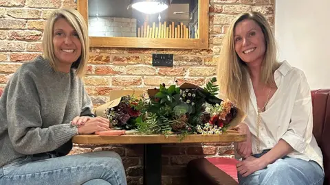 Gemma Kelsall sits at one side of a table across from Sally Lewis, with flowers in the middle. Both have blonde hair and are smiling.