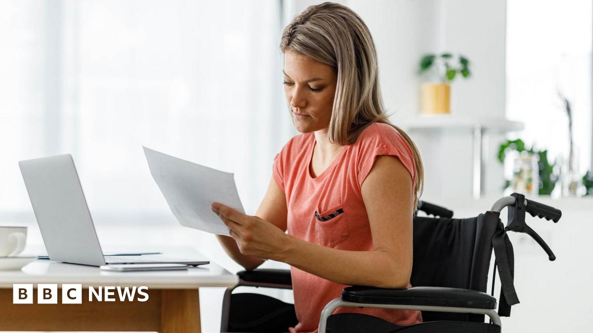 A woman sitting in a wheelchair in front of her laptop, looking at paperwork.