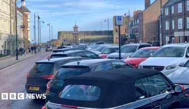 A view down Tynemouth Front Street, with cars parked diagonally in spaces down the middle of the road. The street is lined with three-storey Georgian buildings, some with shop fronts, and ends with a view of a clock tower and old castle walls.