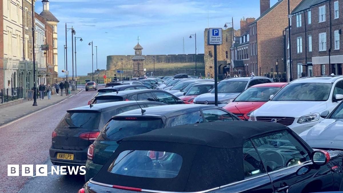 A view down Tynemouth Front Street, with cars parked diagonally in spaces down the middle of the road. The street is lined with three-storey Georgian buildings, some with shop fronts, and ends with a view of a clock tower and old castle walls.