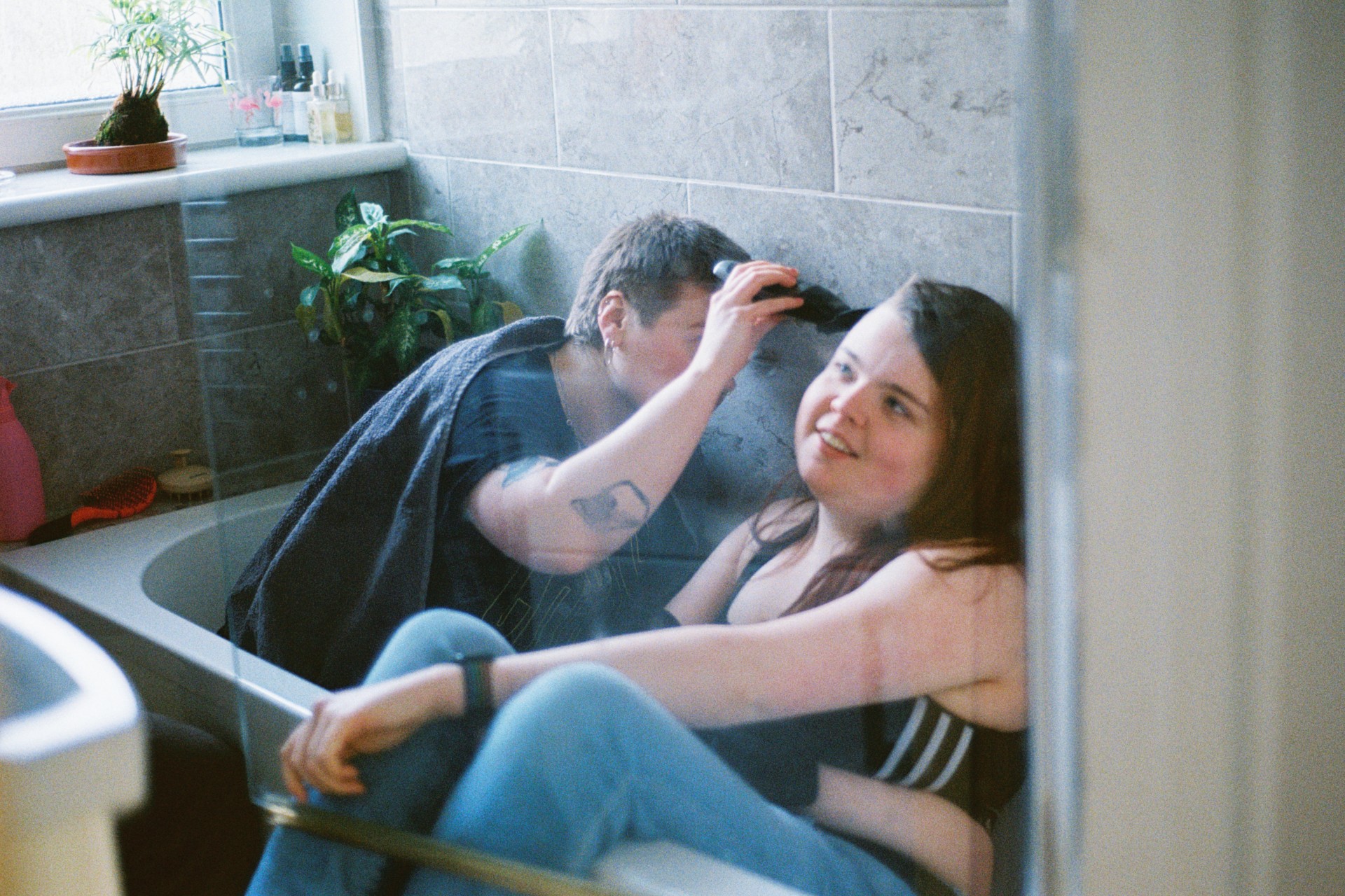 Inside a tiled bathroom, one person sits in a bathtub while another leans over to shave their head with electric clippers.