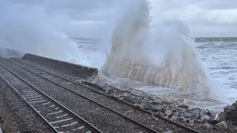 Steve Briars A sea wall is damaged with stones lining the ground.  A large wave is also hitting the wall, sending sea spray into the air.