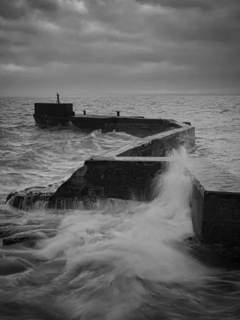 Pete McNaught Black‑and‑white photo of a zigzagging stone pier extending into rough seas. Waves crash against the pier, sending water spilling over the edges. 