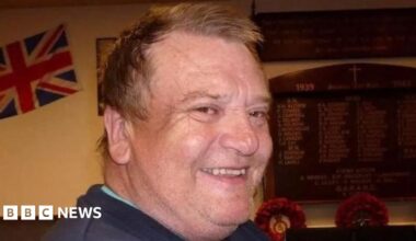 Peter Coates is looking over his shoulder and smiling brightly at the camera. He has short brown hair and is wearing a blue jumper. There is a wooden memorial board on the wall behind him, as well as a union jack flag.