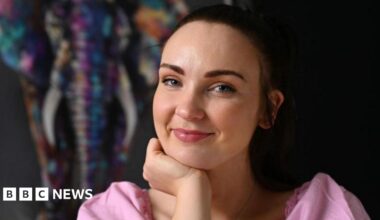 Diona Doherty, with brown hair, tied back. She is wearing a pink top and is holding one hand up to her chin.