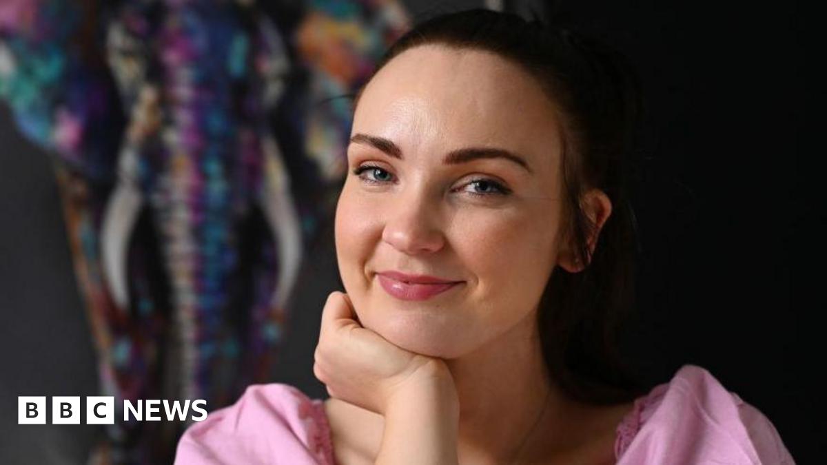 Diona Doherty, with brown hair, tied back. She is wearing a pink top and is holding one hand up to her chin.
