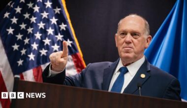 Frey in dark suit and black-and-white tie walks up stairs surrounded by reporters holding their phones out to record him
