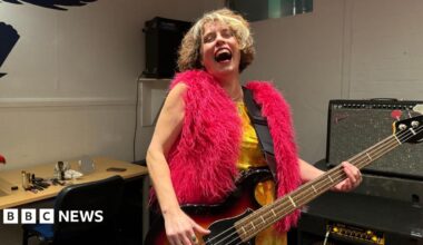 Janet is in a music studio playing her guitar and giving a big laugh with her head back, wearing a bright pink fluffy gilet
