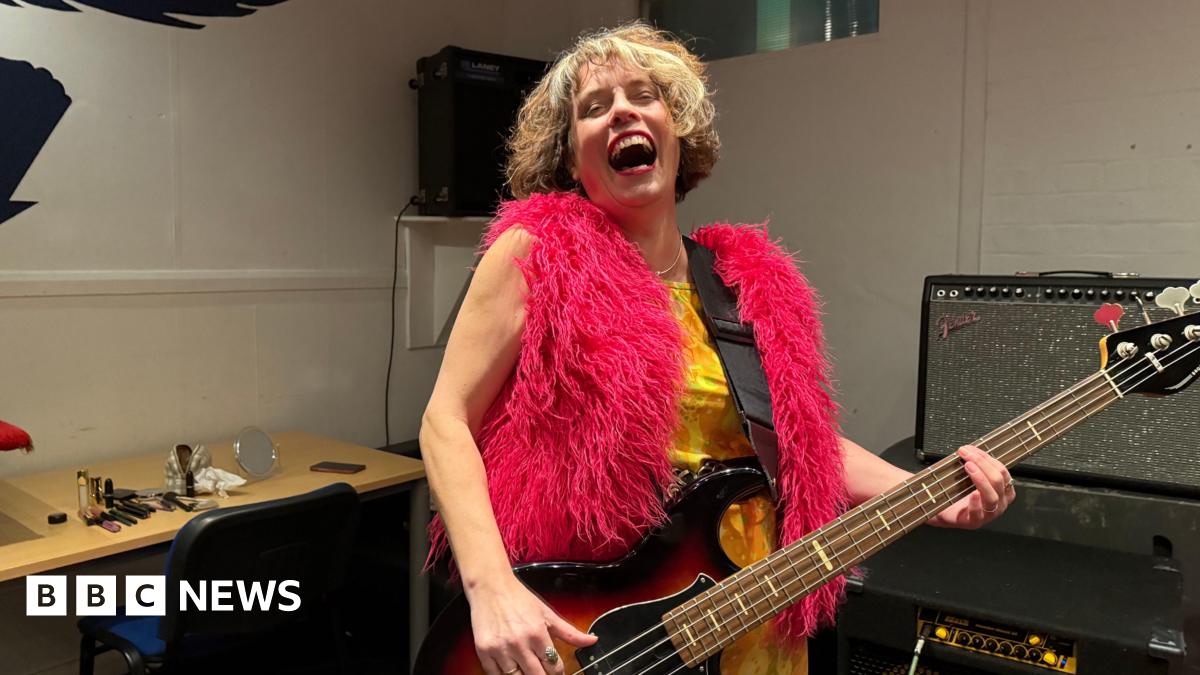 Janet is in a music studio playing her guitar and giving a big laugh with her head back, wearing a bright pink fluffy gilet