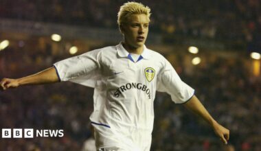 A young Alan Smith celebrating a goal for Leeds United, with his arms outstretched. He has spiky blond hair and the 'Strongbow' logo is prominent on the jersey.